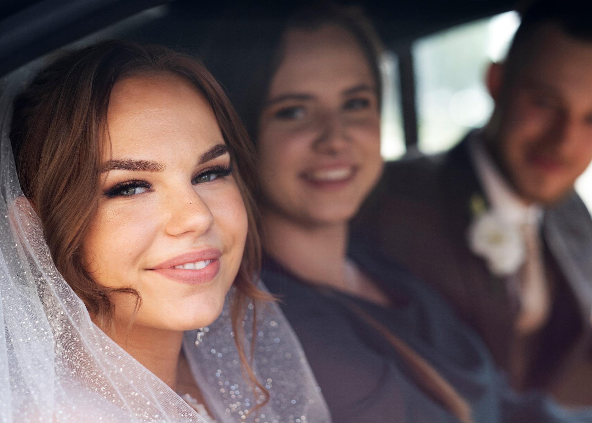 Panna młoda w welonie z przyjaciółką i panem młodym w samochodzie w drodze na ceremonię ślubną, uśmiechnięta, z bliska.