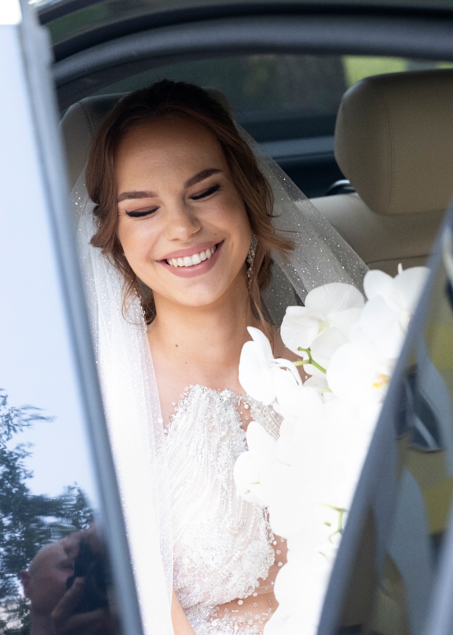 Uśmiechnięta panna młoda w sukni z koronkowymi zdobieniami i welonem, siedząca w samochodzie z bukietem białych orchidei.
