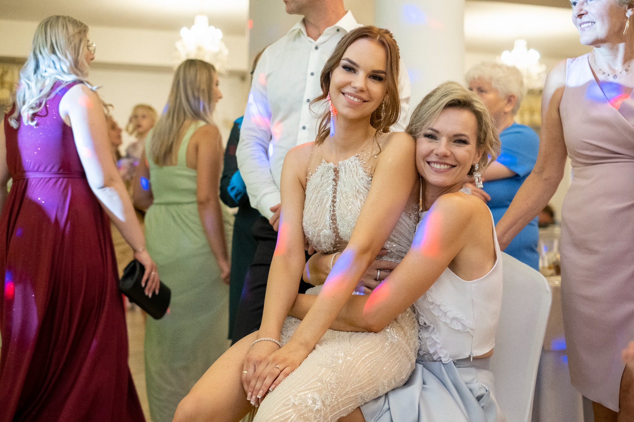 Dwie uśmiechnięte kobiety w sukienkach wieczorowych, siedzące obok siebie na tle gości weselnych i dekoracji świetlnych.