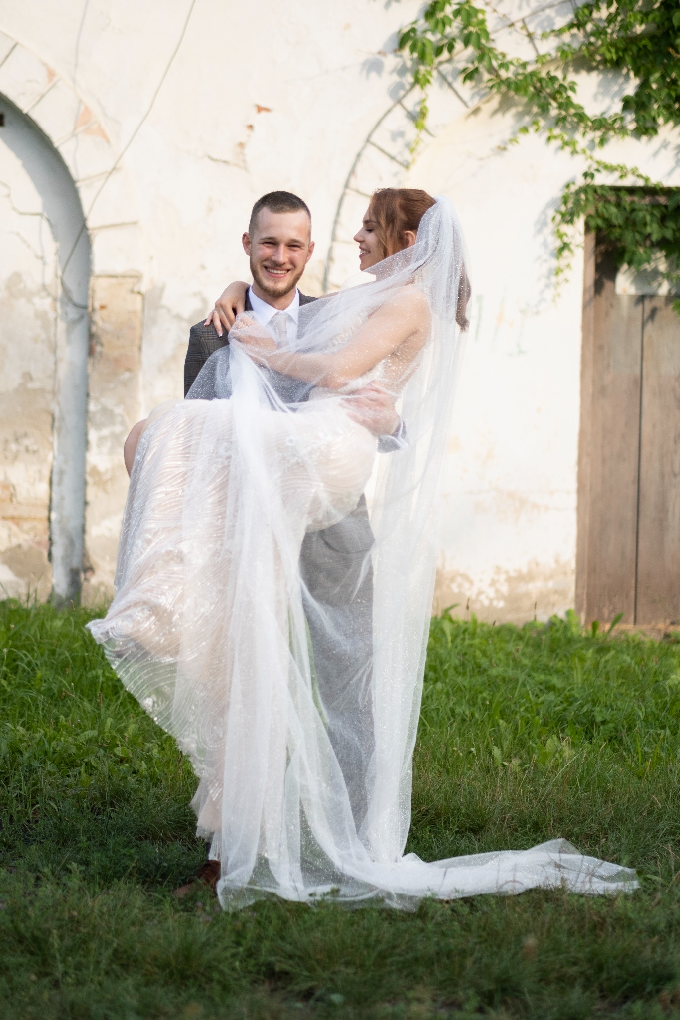 Uśmiechnięty pan młody trzyma na rękach pannę młodą w białej sukni i welonie na tle starej ściany z roślinnością.