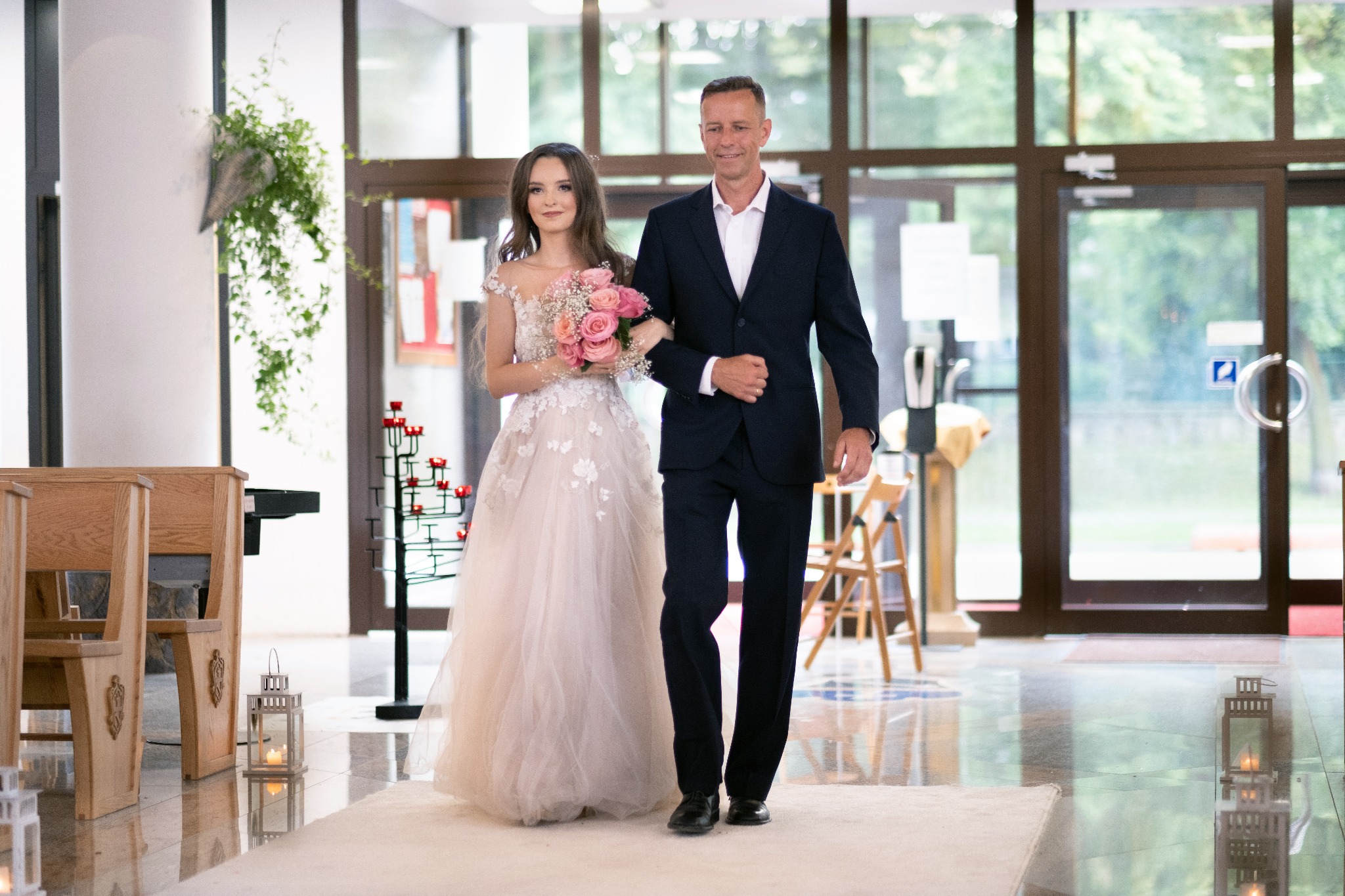 Panna młoda w jasnej sukni ślubnej, prowadzona do ołtarza przez mężczyznę w granatowym garniturze, trzyma bukiet różowych róż, w tle kościelne ławki i dekoracje.