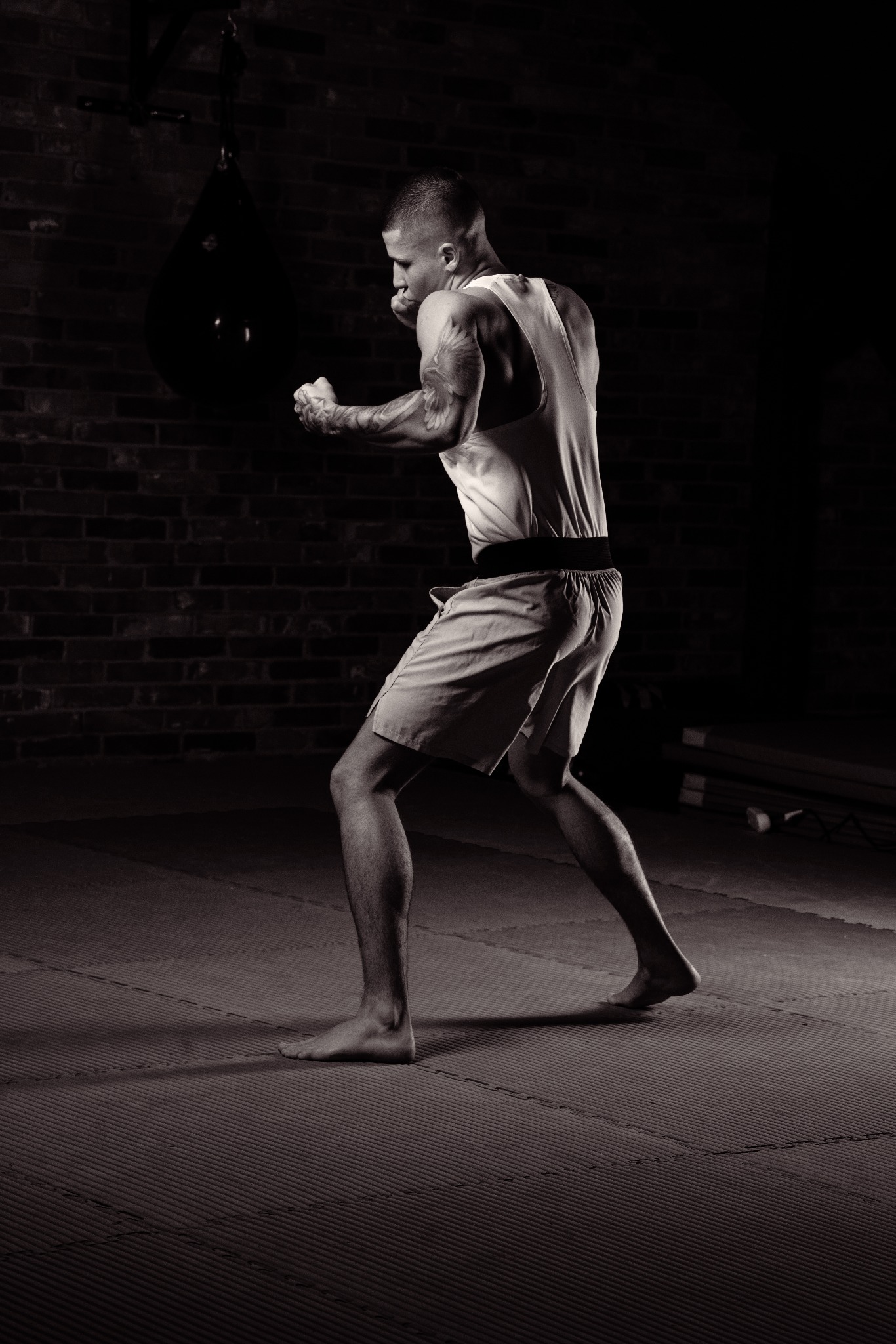 Czarno-białe zdjęcie wytatuowanego mężczyzny w koszulce na ramiączkach i spodenkach bokserskich, trenującego boks na tle ceglanej ściany i wiszącego worka treningowego w zaciemnionym pomieszczeniu,...