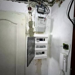 STANEL Usługi elektroinstalacyjne i automatyka budynkowa. Stanisław Okrzesik - Rozdzielnia elektryczna po modyfikacji, widoczny licznik energii, zabezpieczenia oraz otwarte drzwiczki rozdzielni na tle ściany w trakcie remontu.