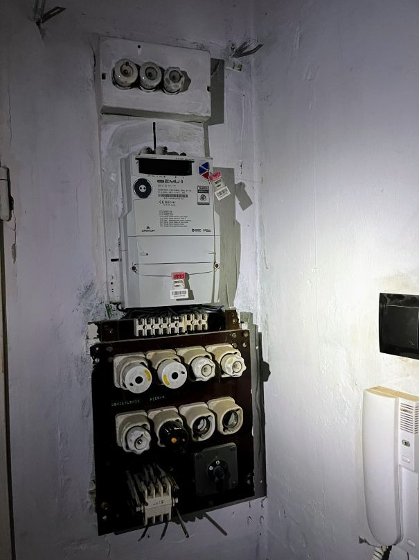 Stara, brudna rozdzielnia elektryczna przed modernizacją, widoczny licznik EMU3, bezpieczniki topikowe i wystające przewody.