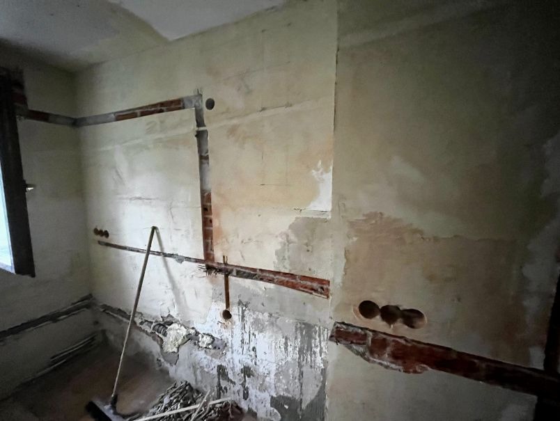 Ściana w trakcie remontu z wyfrezowanymi bruzdami pod kable elektryczne, widoczne otwory pod gniazdka i wystające rurki instalacyjne, zanieczyszczenia na podłodze.