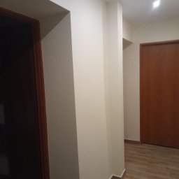 Olexbud Grzegorz Oleksy - Korytarz po odświeżeniu, z jasnymi ścianami, drewnianymi panelami podłogowymi ułożonymi wzdłuż, widoczne drzwi z drewnianą ramą, narożnik ściany z listwą przypodłogową.