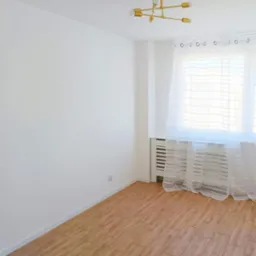 Jasne, puste pomieszczenie z drewnianą podłogą, białymi ścianami, grzejnikiem pod oknem z delikatną firanką i nowoczesną, złotą lampą sufitową.