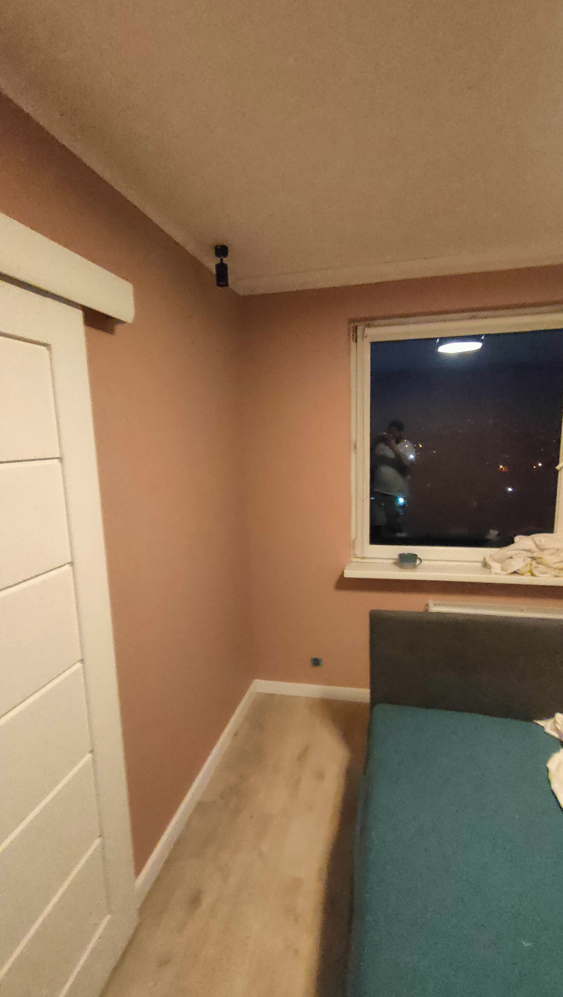 Świeżo pomalowany pokój w odcieniach pudrowego różu po zalaniu, widoczna nowa podłoga z paneli, białe drzwi przesuwne i odbicie osoby w oknie.