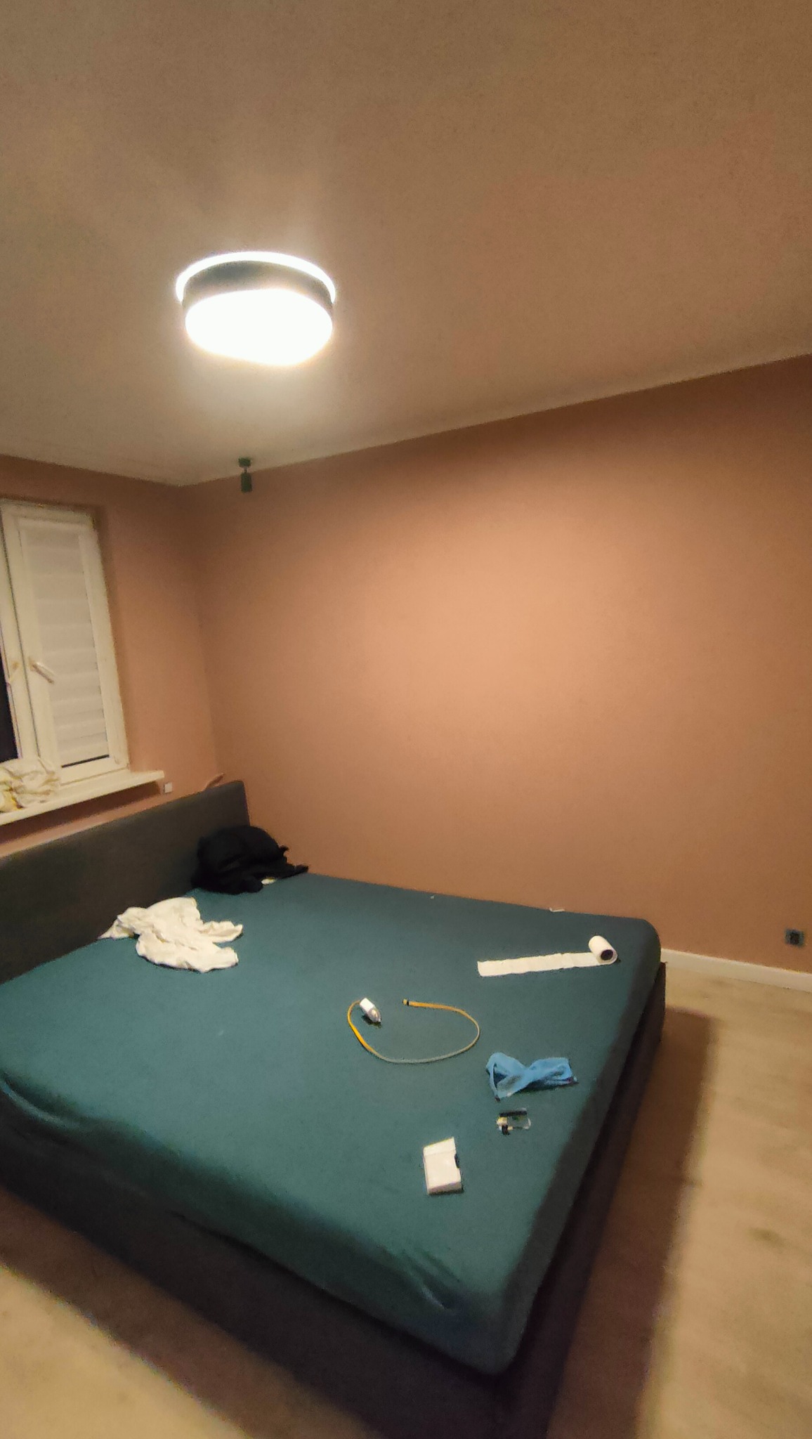 Świeżo pomalowany pokój z widocznymi smugami na ścianie i rozrzuconymi przedmiotami na łóżku, w tym wałek do malowania i taśma malarska.