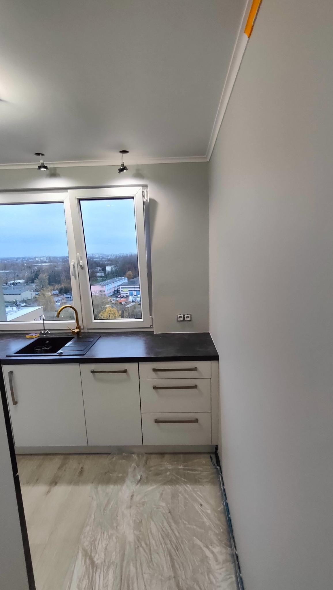 Świeżo pomalowana kuchnia w szarościach, z białymi szafkami, czarnym blatem i złotą armaturą, podłoga zabezpieczona folią malarską.