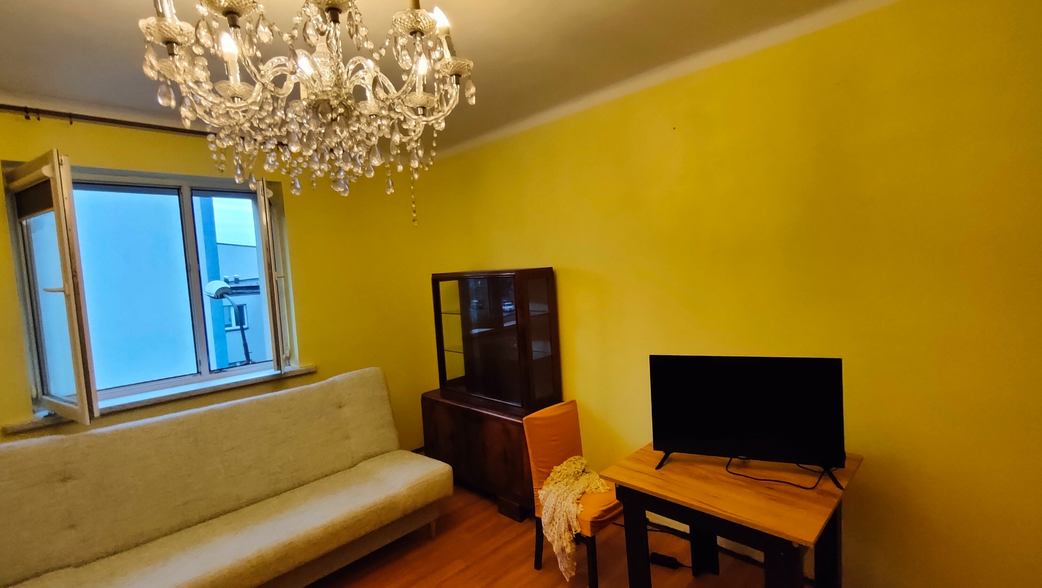 Żółty pokój z otwartym oknem, kryształowym żyrandolem, sofą, komodą, krzesłem z narzutą i telewizorem na drewnianym stoliku.