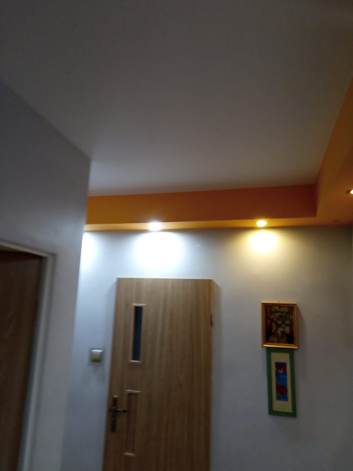 Wnętrze korytarza po remoncie: świeżo pomalowany sufit z pomarańczowym akcentem, zamontowane drzwi wewnętrzne z wąskim przeszkleniem i dekoracje ścienne w postaci dwóch obrazków.