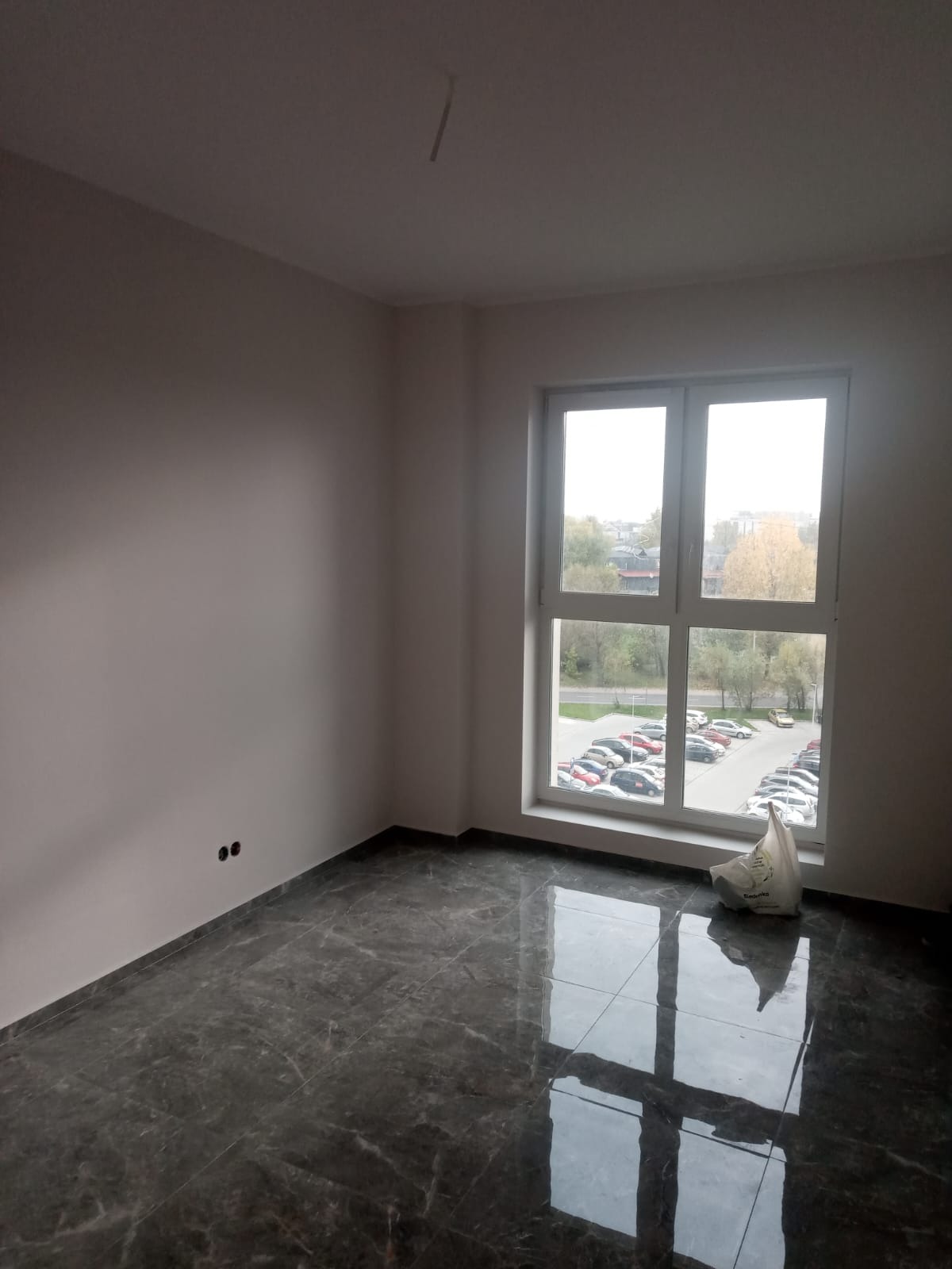 Pusty pokój po remoncie z ciemną, błyszczącą podłogą imitującą marmur, świeżo pomalowane ściany w odcieniach szarości, widoczne dwa gniazdka elektryczne na ścianie oraz okno z widokiem na parking.