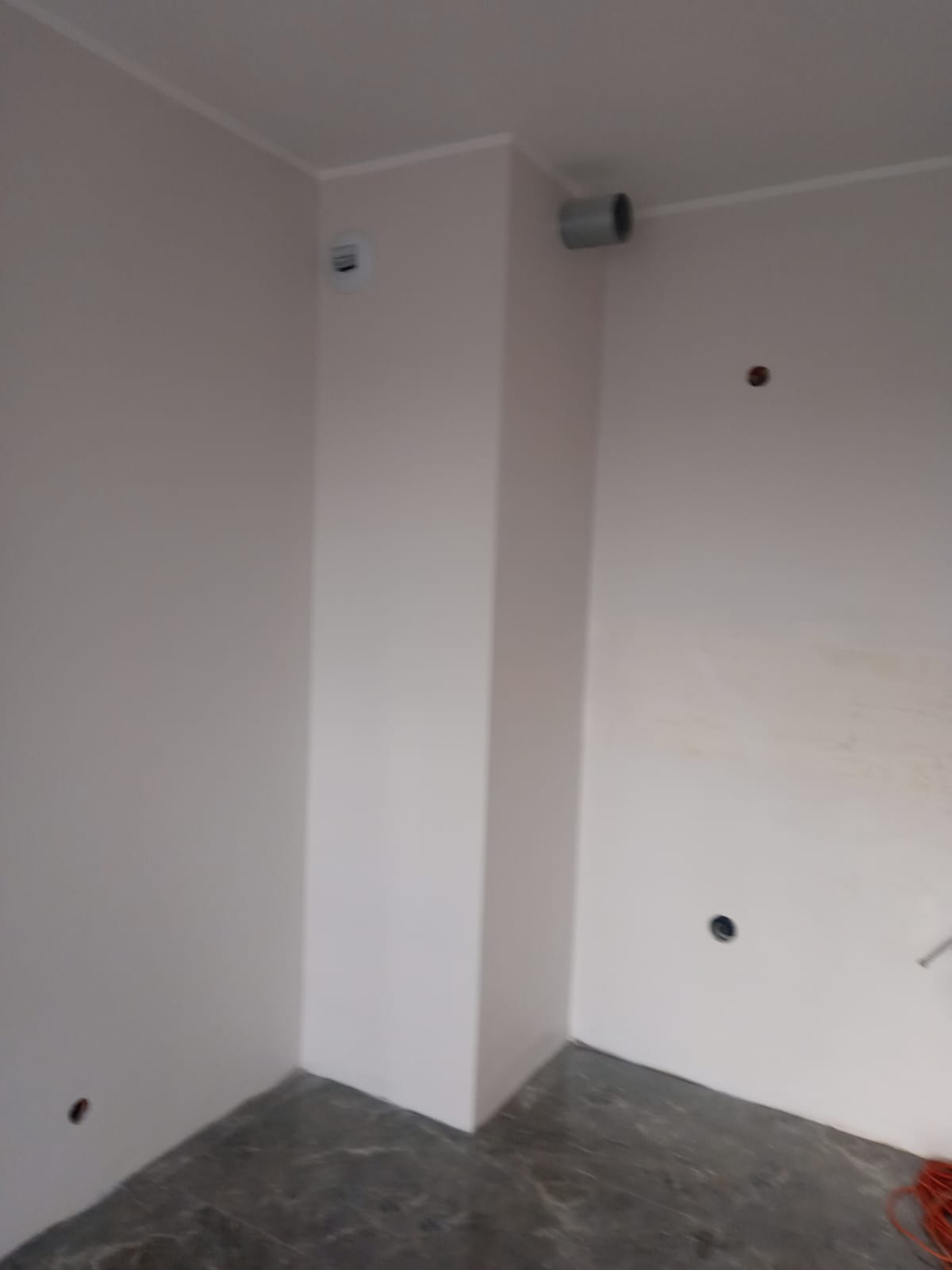 Wnętrze po świeżym pomalowaniu ścian na jasny odcień, widoczne gniazdka elektryczne i rura wentylacyjna przy suficie, szara podłoga z wzorem marmuru.