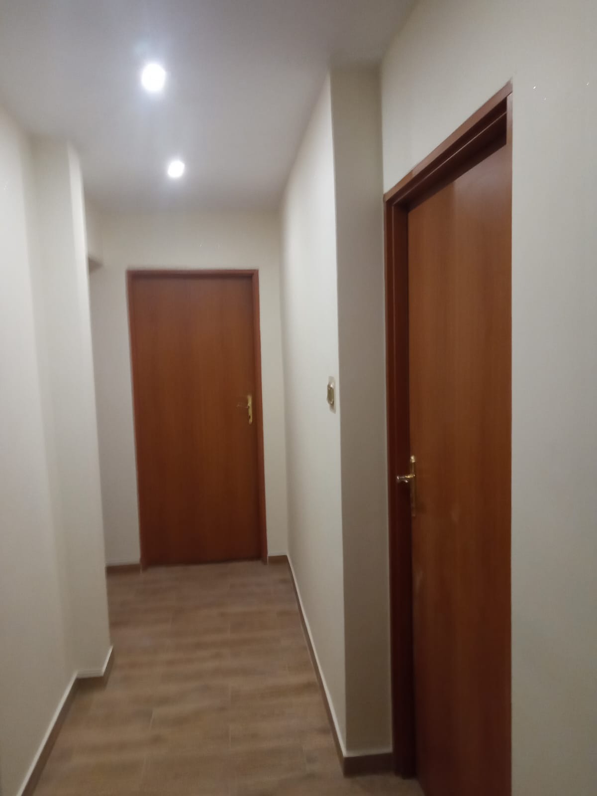 Korytarz po remoncie z dwiema drewnianymi drzwiami i podłogą imitującą drewno, oświetlony punktowymi lampami sufitowymi.