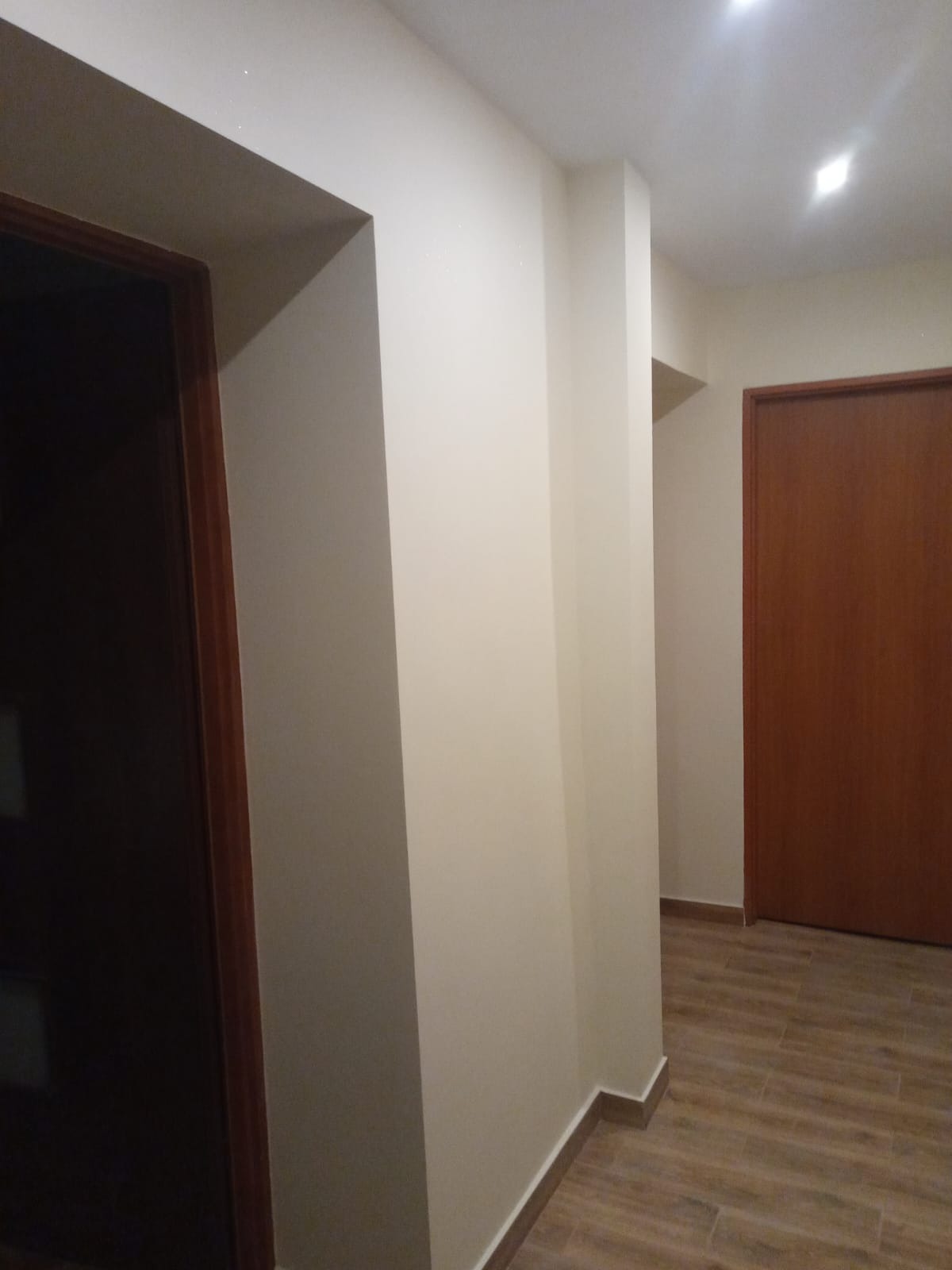 Korytarz po odświeżeniu, z jasnymi ścianami, drewnianymi panelami podłogowymi ułożonymi wzdłuż, widoczne drzwi z drewnianą ramą, narożnik ściany z listwą przypodłogową.