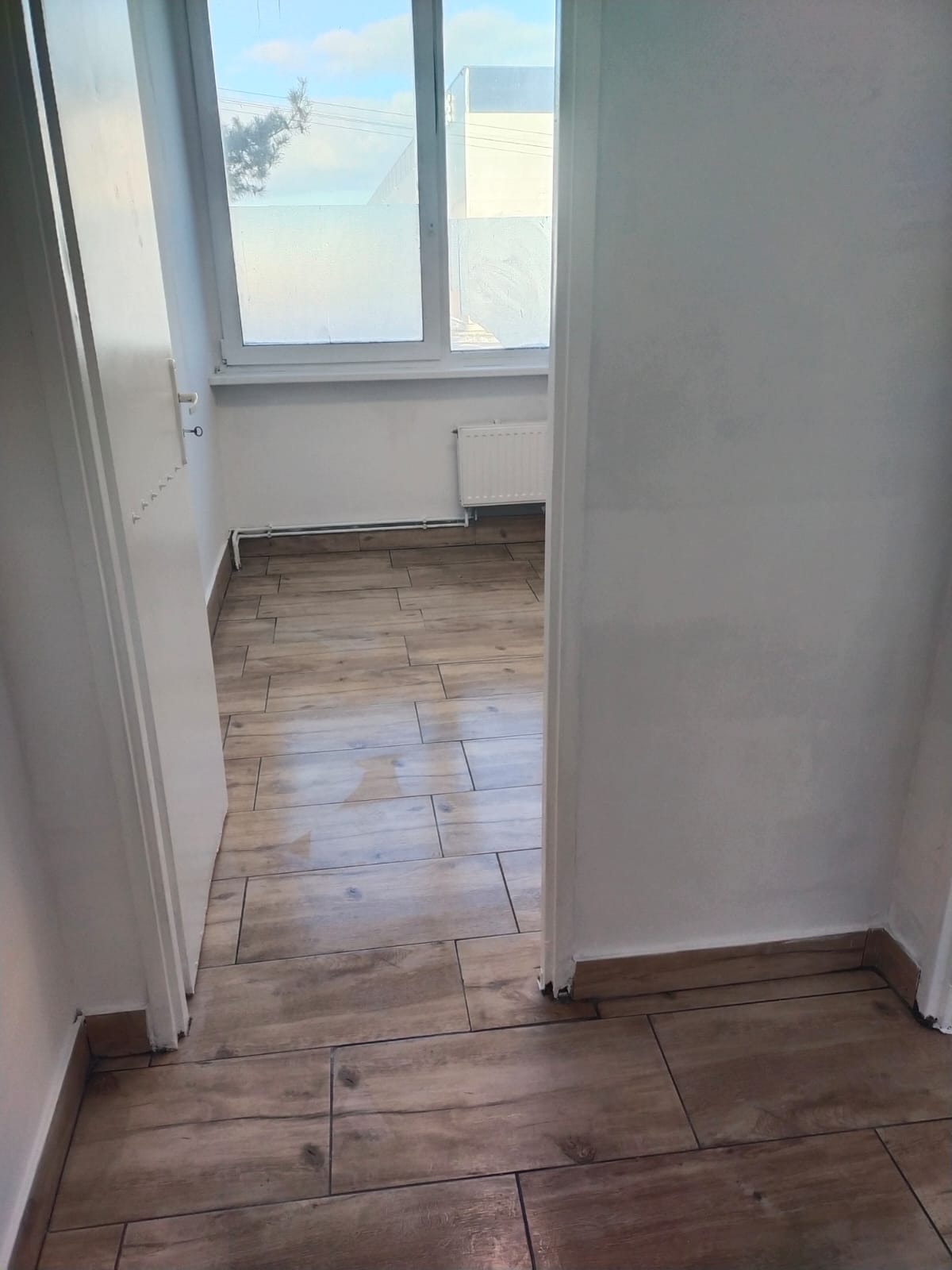 Widok z korytarza na pokój z ułożonymi panelami podłogowymi imitującymi drewno, białe ściany i okno z mleczną szybą.