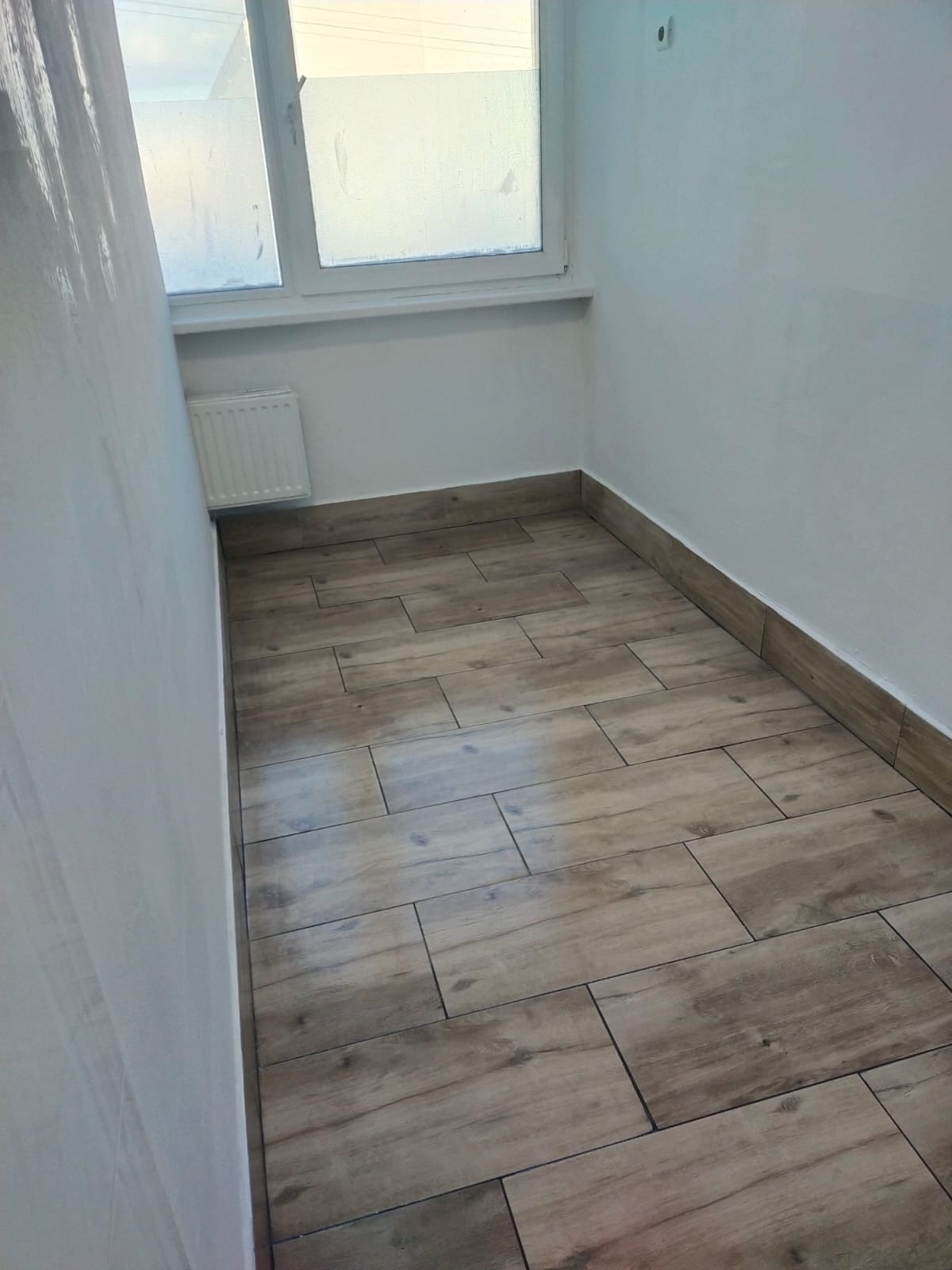 Ułożone panele podłogowe imitujące drewno w małym pomieszczeniu z grzejnikiem pod oknem i białymi ścianami.