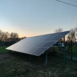 Instalacja fotowoltaiczna na gruncie, widok z boku o zmierzchu, panele ustawione pod kątem na metalowych wspornikach, w tle drzewa i ogrodzenie.