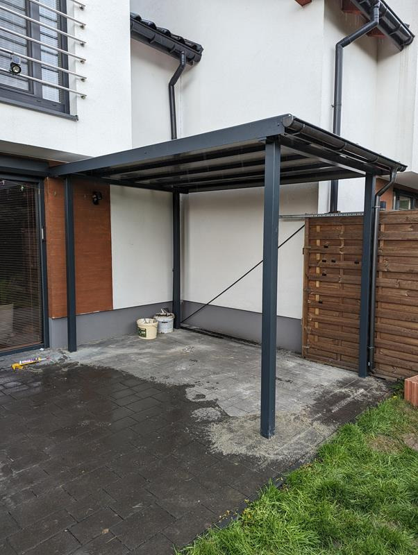 Szara metalowa pergola przy białym budynku z ciemną stolarką okienną, widoczne dwa wiadra i ślady po pracach budowlanych na kostce brukowej.