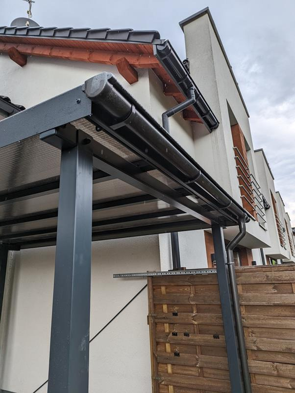 Szara metalowa pergola przylegająca do budynku z drewnianym ogrodzeniem w tle, widok z dołu na konstrukcję i rynny.