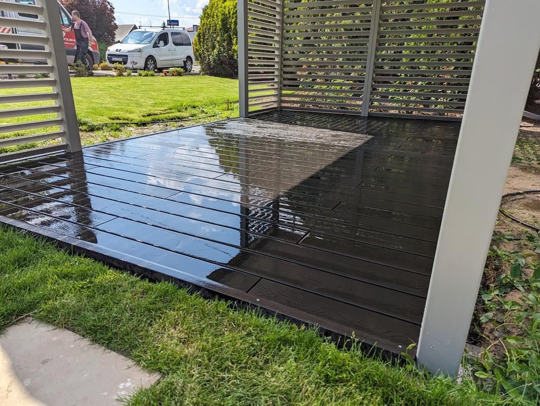 Nowoczesny taras z desek kompozytowych w kolorze ciemnego brązu, zadaszony metalową pergolą z poziomymi lamelami, świeżo po umyciu, z widocznymi odbiciami nieba na mokrej powierzchni.
