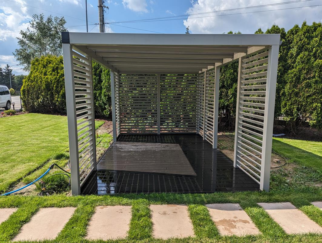 Nowoczesna, metalowa pergola w kolorze szarym, z lamelami, na ciemnym tarasie kompozytowym, otoczona zielonym trawnikiem i ścieżką z płyt chodnikowych. Na trawniku widoczny zwinięty wąż ogrodowy.