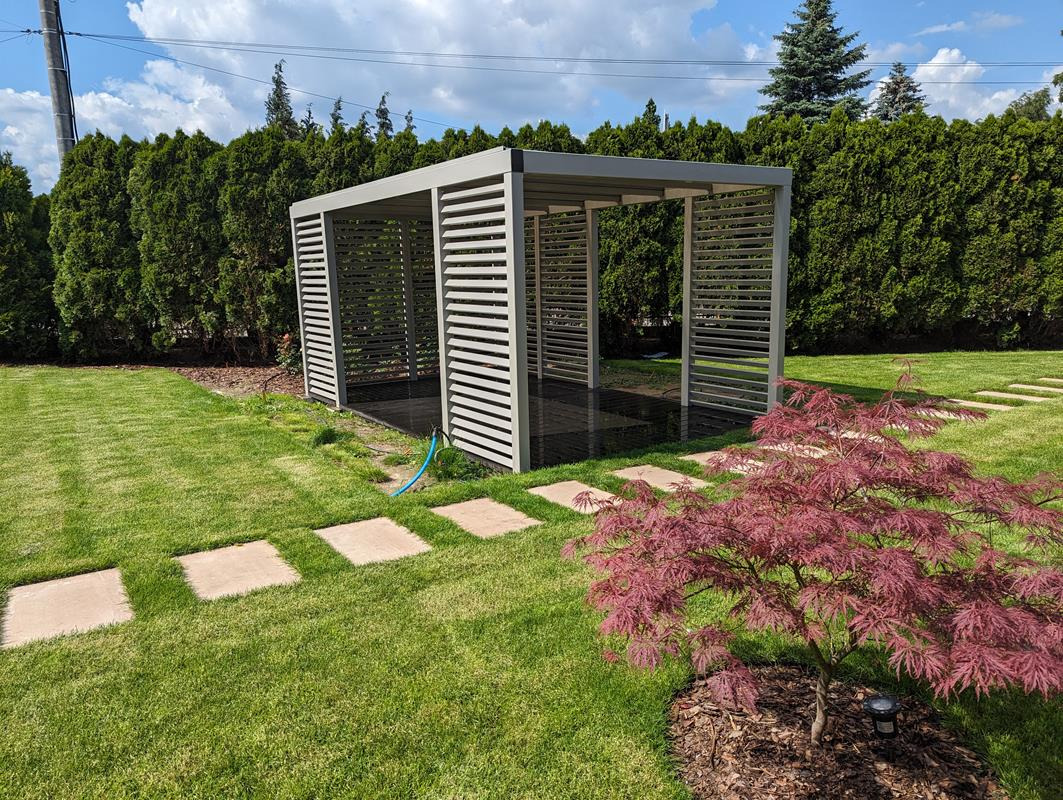 Nowoczesna pergola metalowa z poziomymi lamelami w ogrodzie w Rzeszowie, widok z perspektywy niskiego, czerwonego klonu japońskiego, otoczona zielonym trawnikiem i tujami.