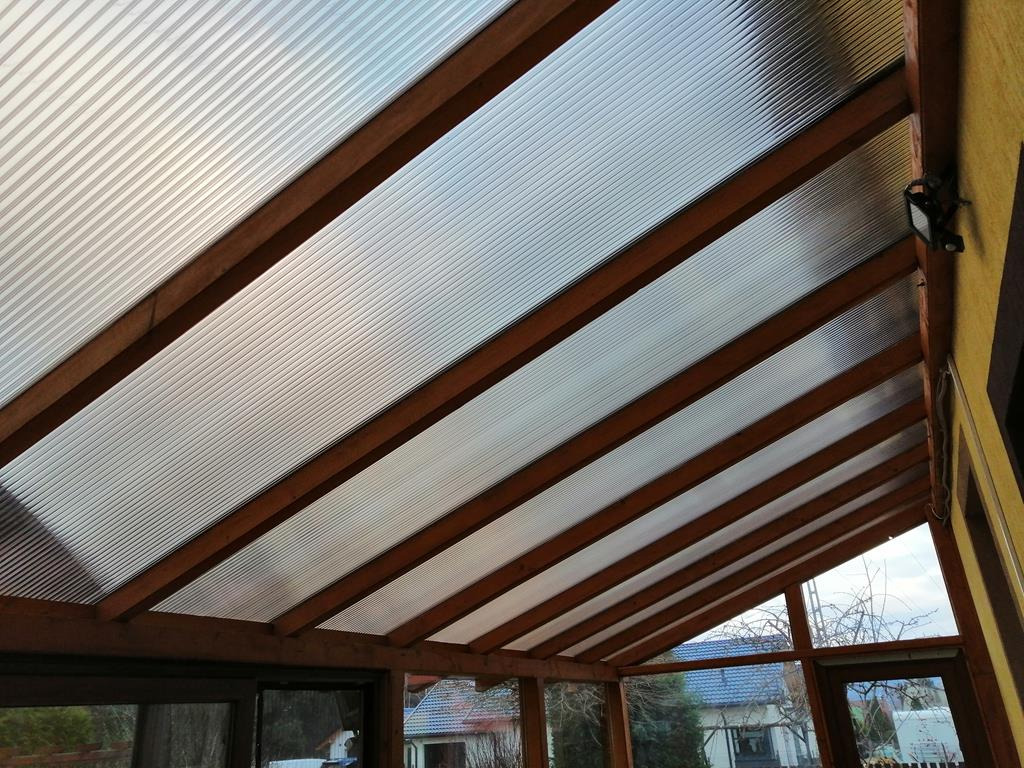 Zadaszenie tarasu z brązowymi drewnianymi belkami i półprzezroczystymi panelami, widok od dołu, z widocznym oświetleniem zewnętrznym na ścianie i fragmentem krajobrazu za oknem.