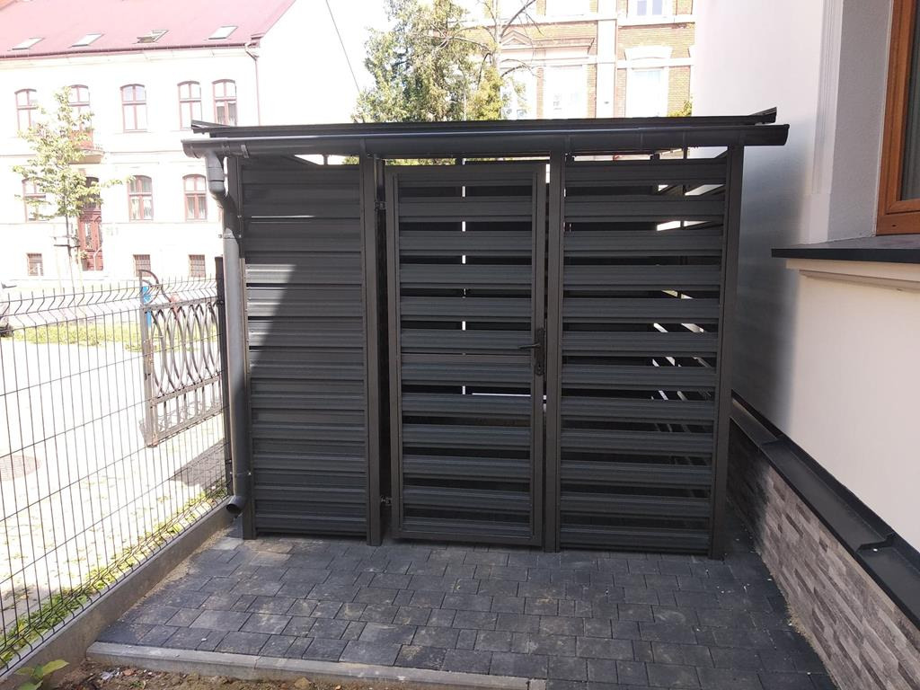 Zamykana, metalowa konstrukcja na śmietnik z poziomymi panelami, umieszczona na brukowanym podłożu, obok budynku.