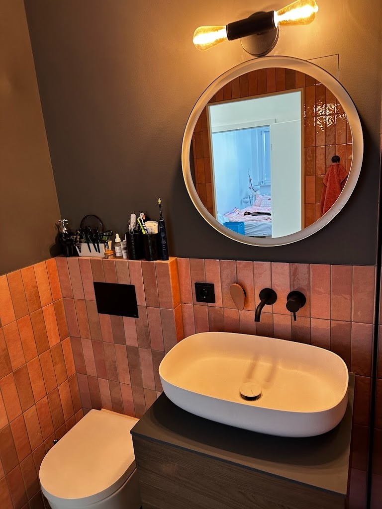 Nowoczesna łazienka z owalną umywalką nablatową, terakotową glazurą ułożoną pionowo, okrągłym lustrem z podświetleniem LED i widoczną w tle sypialnią.