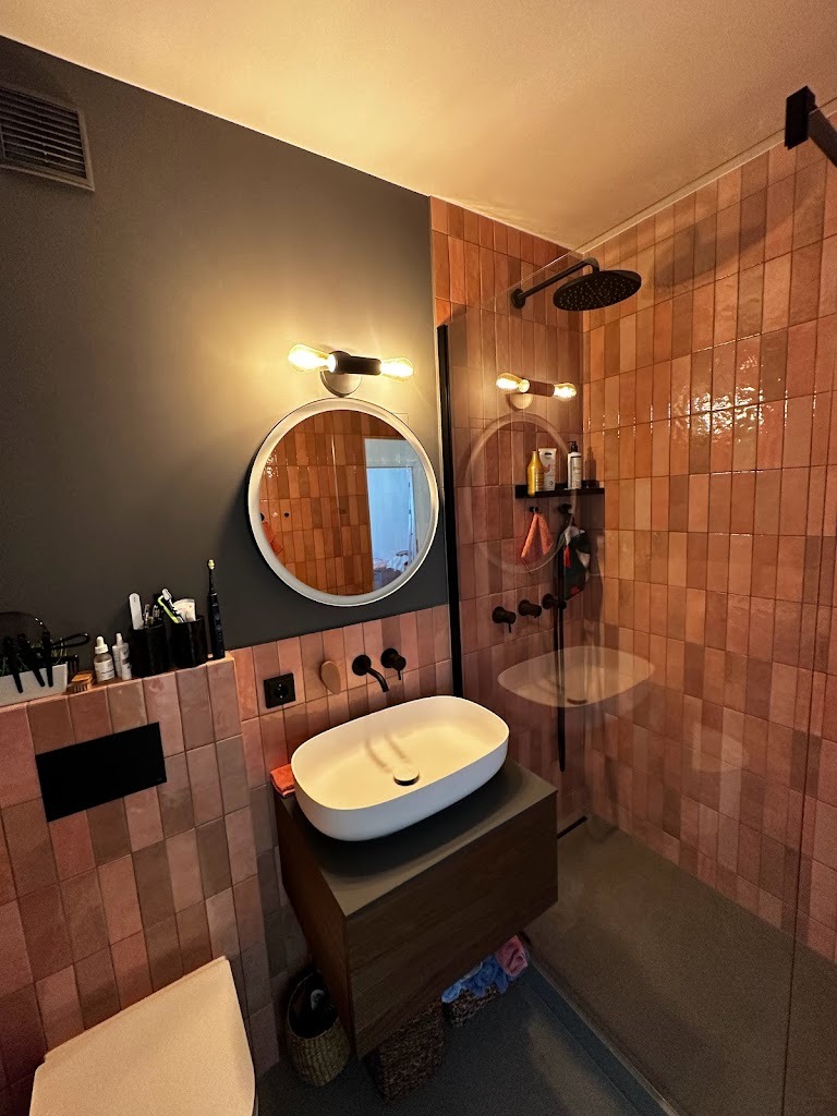 Nowoczesna łazienka z terakotową glazurą, umywalką nablatową na drewnianej szafce i czarną armaturą, oświetlona ciepłym światłem.