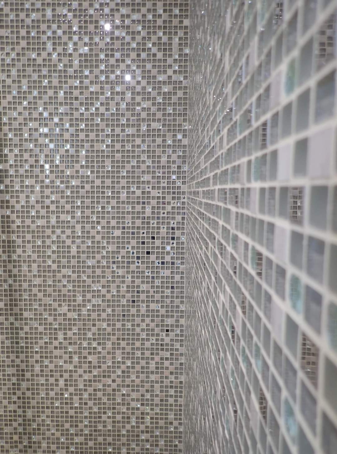 Ściana wyłożona mozaiką z drobnych, kwadratowych płytek w odcieniach szarości, z błyszczącymi, szklanymi elementami odbijającymi światło, widoczna pod kątem, ukazująca narożnik pomieszczenia.