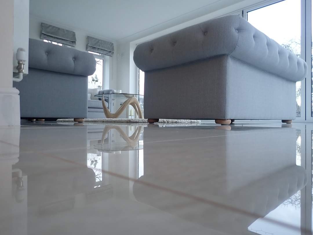 Błyszcząca, jasna podłoga z płytek odbija fragmenty szarych mebli tapicerowanych i stolika kawowego o szklanym blacie w minimalistycznym, nowoczesnym salonie z dużymi oknami.