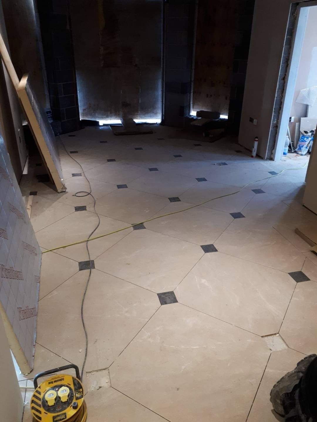 Podłoga z jasnych płytek z ciemnymi wstawkami w trakcie układania, widoczna żółta przedłużka kablowa i płyty izolacyjne opierające się o ścianę, surowy stan ścian w tle.