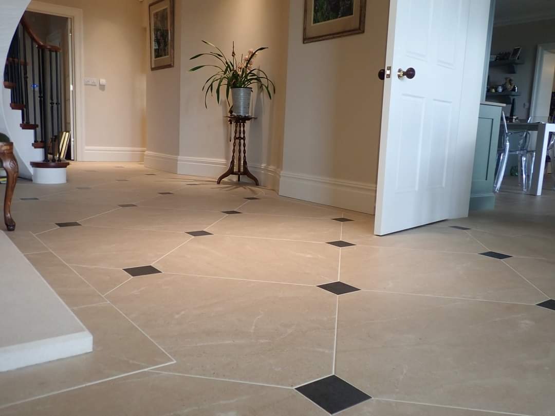 Beżowa podłoga z płytek o nieregularnym kształcie z ciemnymi, kwadratowymi wstawkami, tworząca geometryczny wzór w przestronnym przedpokoju z otwartymi drzwiami i elementami dekoracyjnymi.