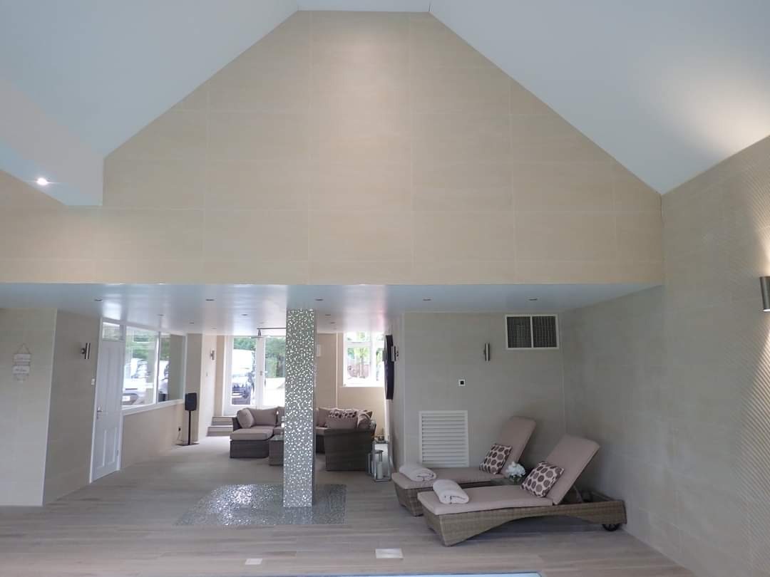 Jasne, przestronne wnętrze z beżowymi płytkami na ścianach i podłodze, mozaikową kolumną, leżakami z poduszkami i widokiem na salon przez przeszklone drzwi.