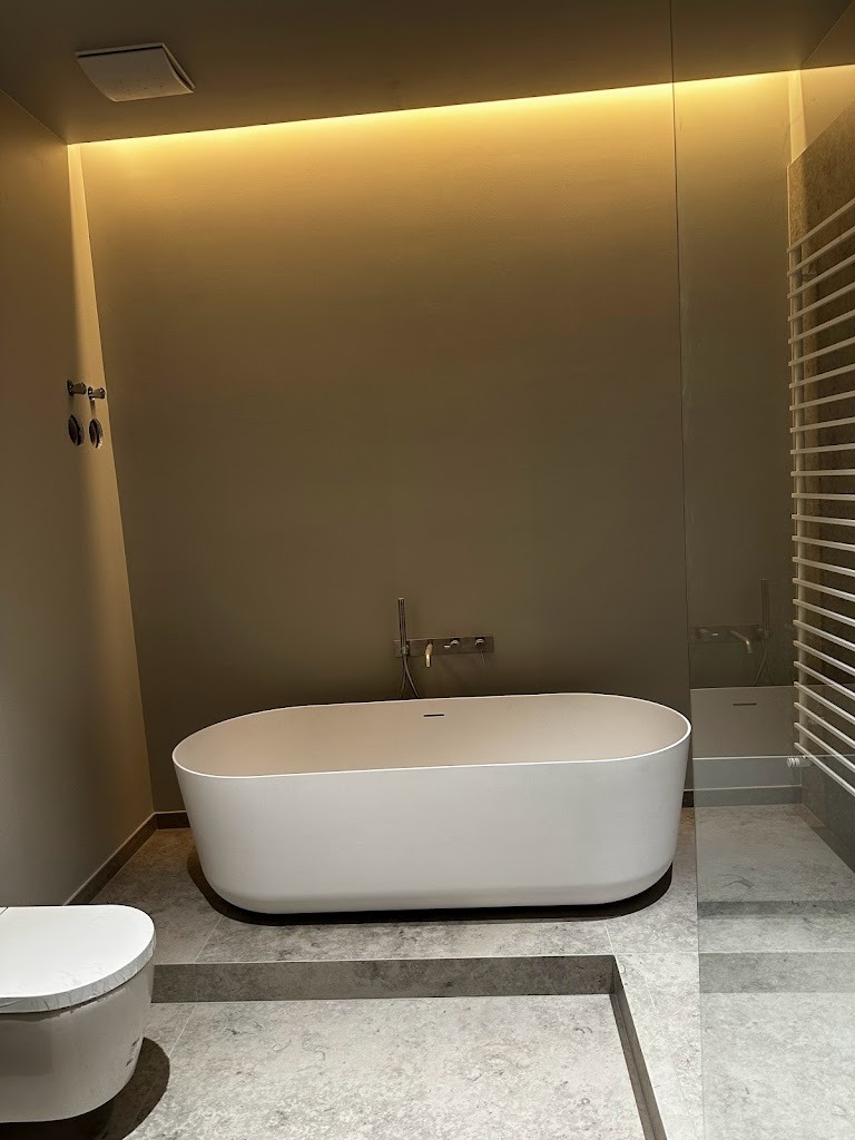 Nowoczesna łazienka z wolnostojącą białą wanną na podwyższeniu z szarego kamienia, minimalistyczna armatura, podświetlenie LED w suficie i grzejnik drabinkowy