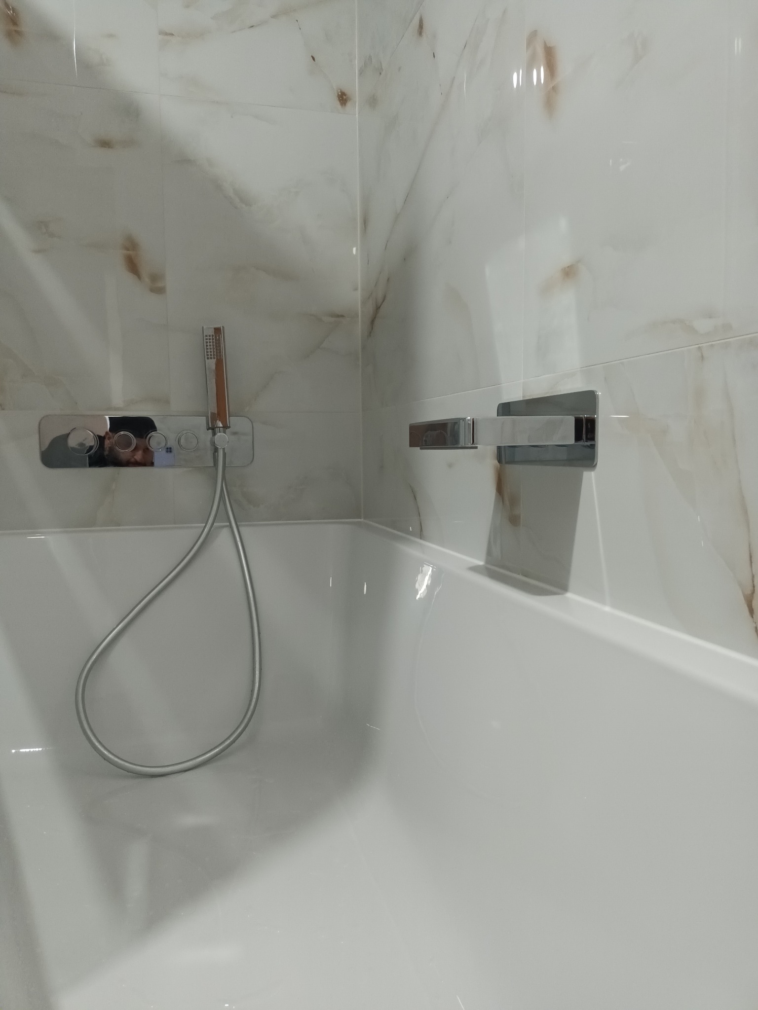 Biała wanna z zamontowaną baterią i prysznicem na tle marmurowych płytek z brązowymi żyłkami.