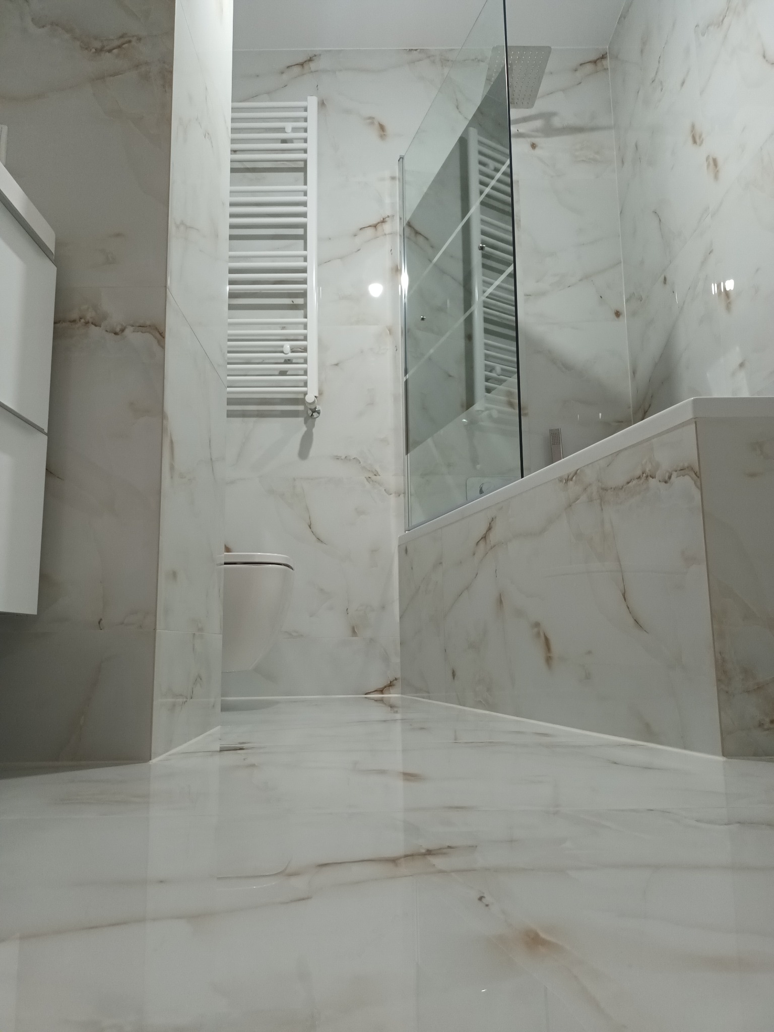 Nowoczesna łazienka wykończona białymi płytkami imitującymi marmur, z grzejnikiem drabinkowym, podwieszaną toaletą i szklanym parawanem nawannowym.