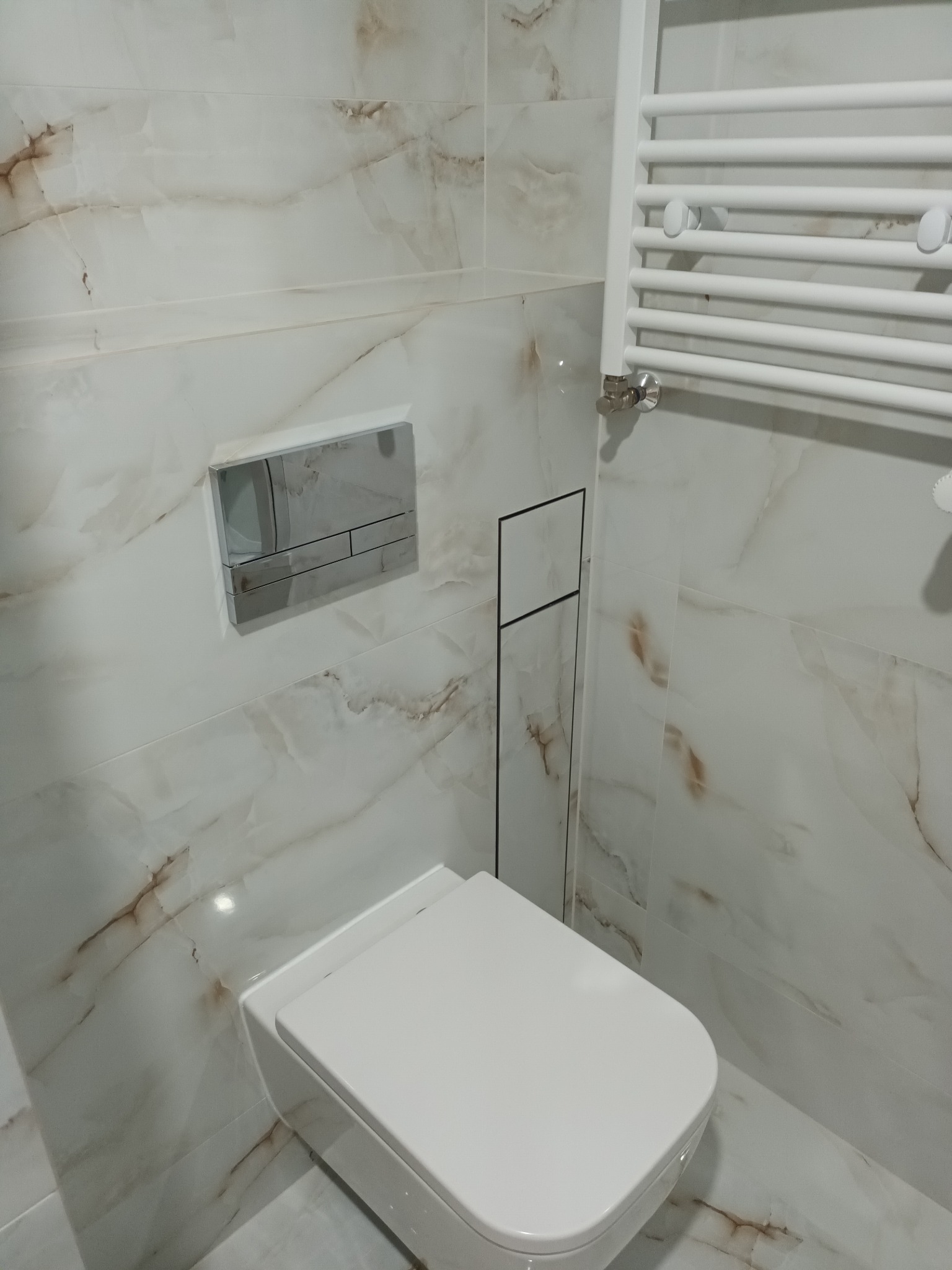 Nowoczesna łazienka z marmurowymi płytkami, podwieszanym WC z chromowanym przyciskiem spłukiwania i białym grzejnikiem drabinkowym.