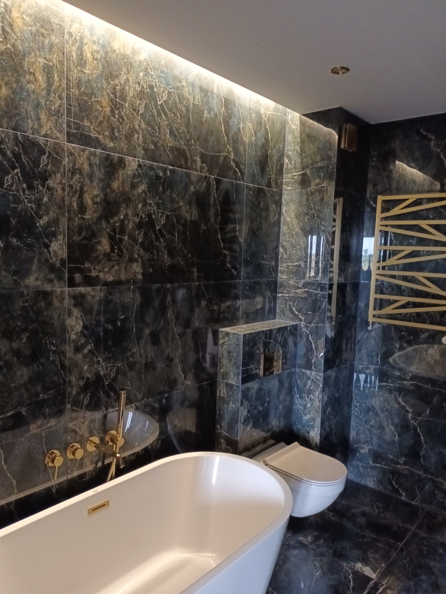 Nowoczesna łazienka wykończona czarnymi płytkami imitującymi marmur, z białą wanną wolnostojącą i złotą armaturą oraz grzejnikiem drabinkowym.
