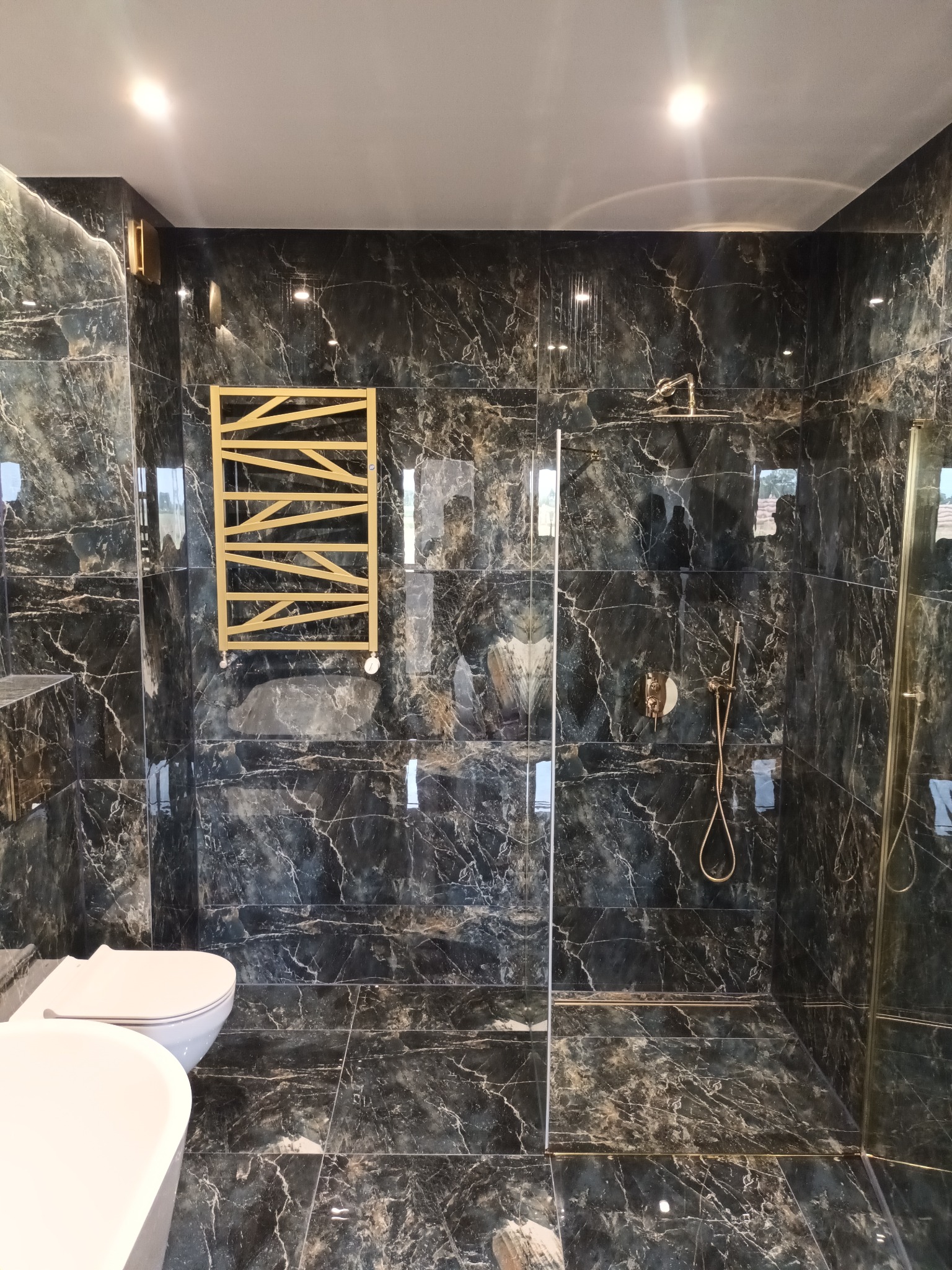 Nowoczesna łazienka z czarnymi płytkami imitującymi marmur, złota grzejnikiem drabinkowym i armaturą w tym samym kolorze, szklana kabina prysznicowa, biała toaleta i fragment wanny.