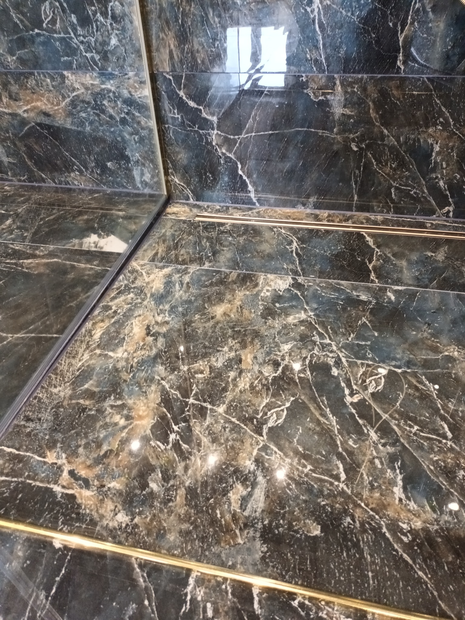 Ciemne, marmurowe płytki na ścianie z delikatnymi złotymi listwami dekoracyjnymi, widoczne odbicie światła na powierzchni płytek.