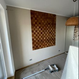 Jakub Ziemiański - Pokój w trakcie malowania na neutralny kolor, zabezpieczona folią podłoga, widoczny wałek w kuwecie i puszka farby, dekoracyjna drewniana mozaika na ścianie.