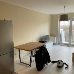 Jakub Ziemiański - Jasne, świeżo pomalowane wnętrze mieszkania z drewnianym stołem, krzesłami i sprzętem AGD. Kremowe ściany i biała podłoga. Minimalistyczny styl aranżacji.