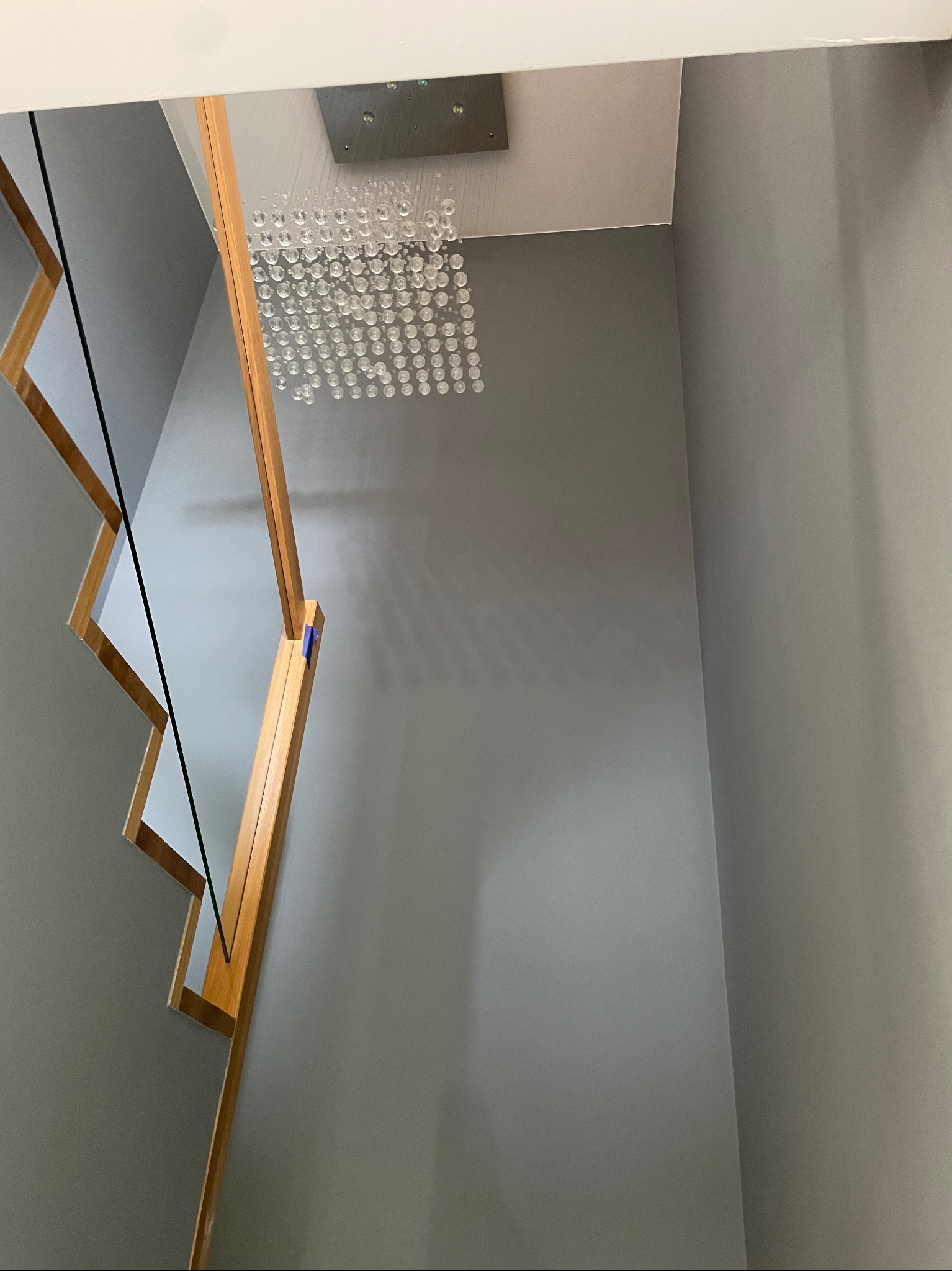 Klatka schodowa z drewnianymi schodami, szklaną balustradą i nowoczesnym żyrandolem na tle szarej ściany.