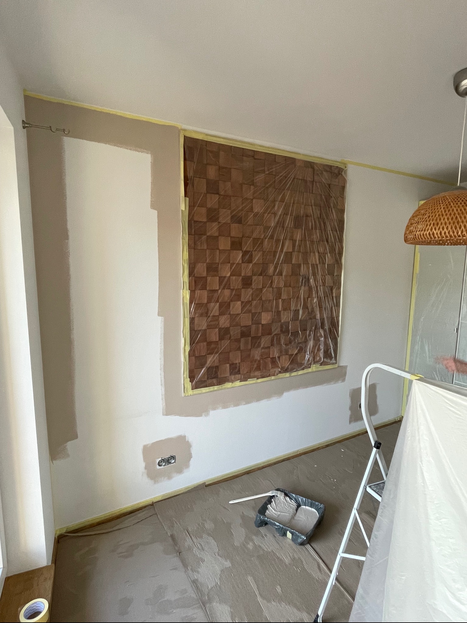 Pokój w trakcie odświeżania koloru ścian, częściowo pomalowane fragmenty w odcieniach beżu, zabezpieczone folią okno z dekoracyjną okładziną, widoczna kuweta z farbą i drabina malarska.
