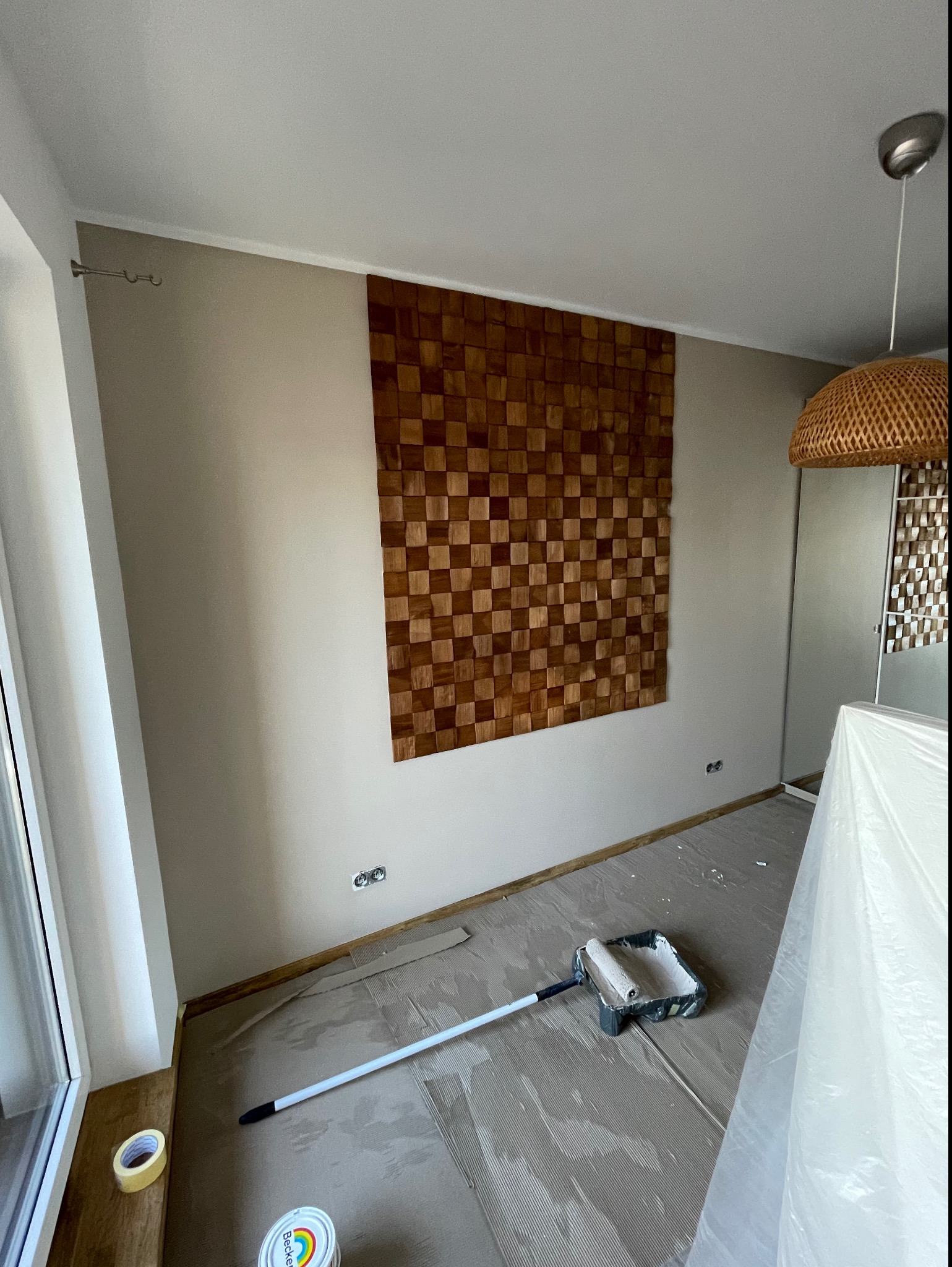 Pokój w trakcie malowania na neutralny kolor, zabezpieczona folią podłoga, widoczny wałek w kuwecie i puszka farby, dekoracyjna drewniana mozaika na ścianie.