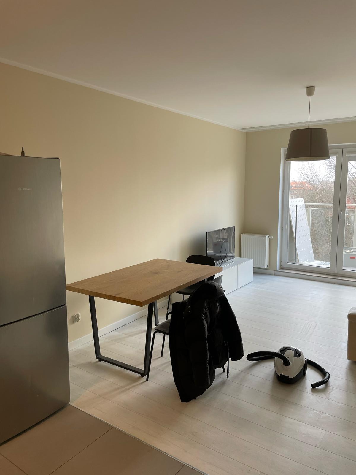 Jasne, świeżo pomalowane wnętrze mieszkania z drewnianym stołem, krzesłami i sprzętem AGD. Kremowe ściany i biała podłoga. Minimalistyczny styl aranżacji.